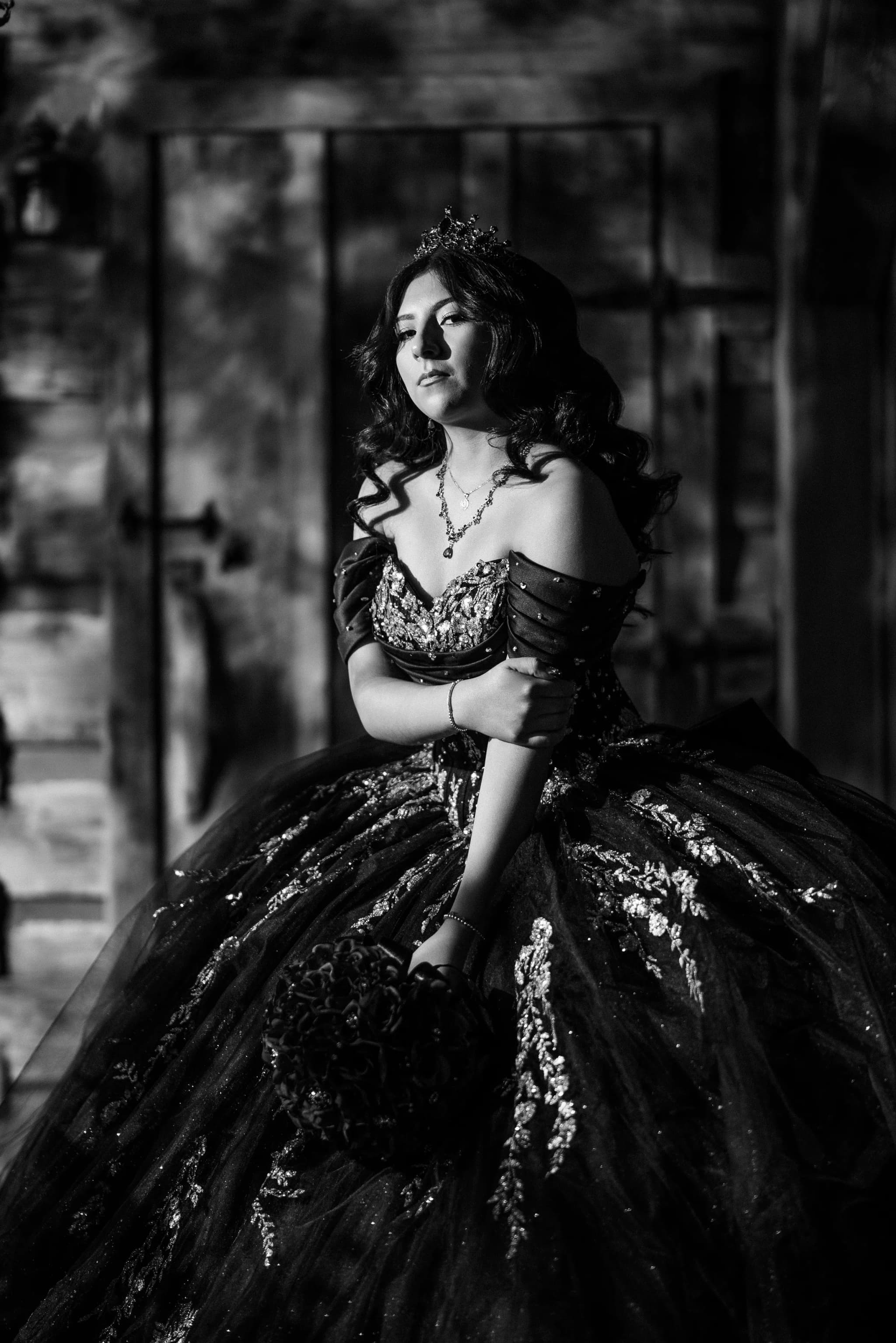 Black-and-white quinceañera portrait in a crowned ball gown before a rustic barn door