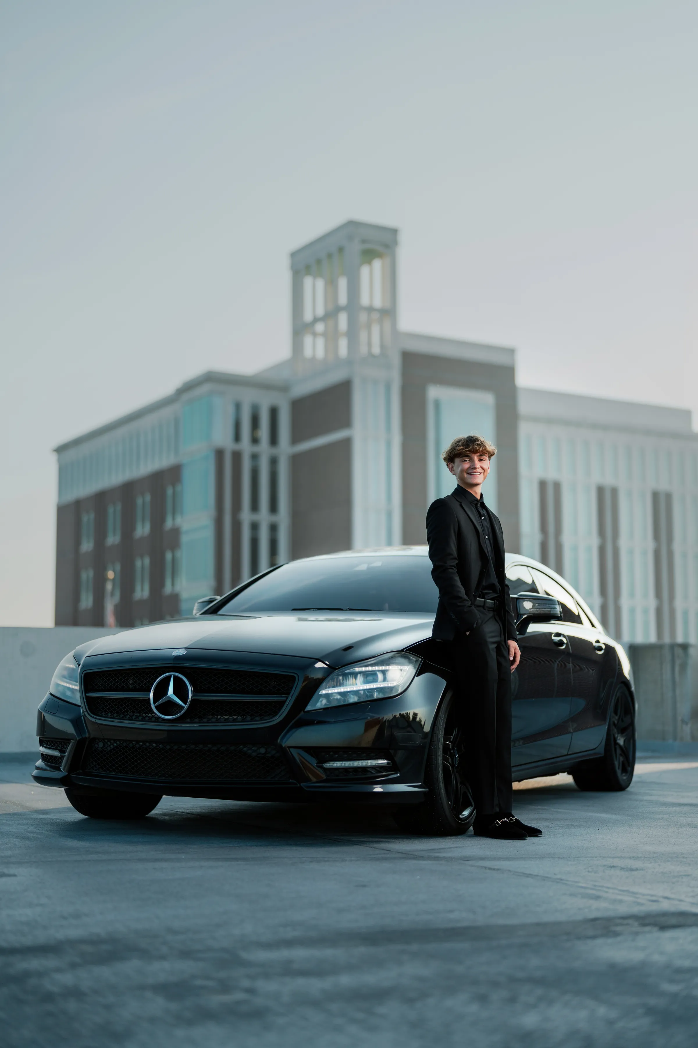 Senior standing beside his black Mercedes on a rooftop deck with a modern building behind
