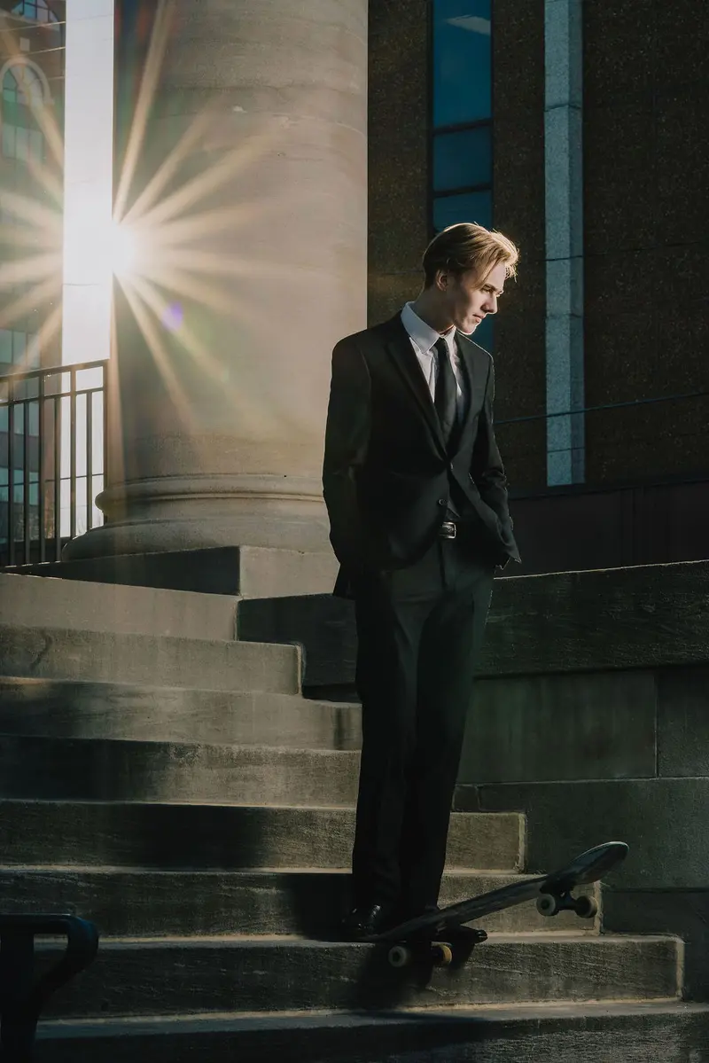 Senior in a black suit resting one foot on a skateboard on courthouse steps with sunburst light flare