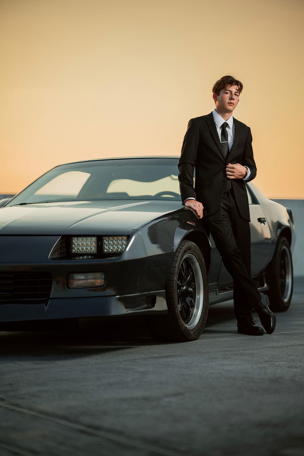 Senior in a black suit leaning on a classic Camaro at sunset