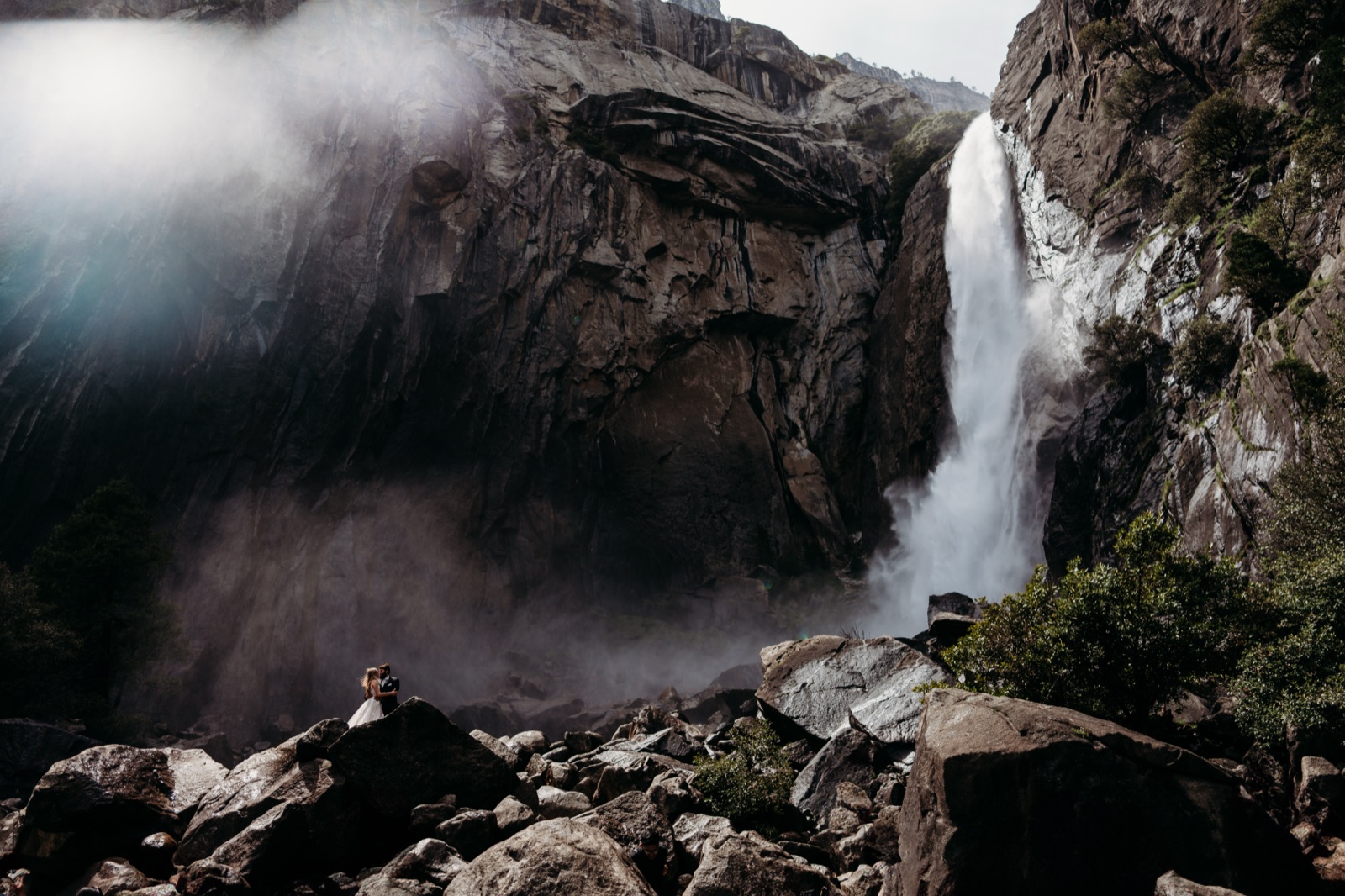 Couple embracing on the rocks beneath a towering Yosemite waterfall
