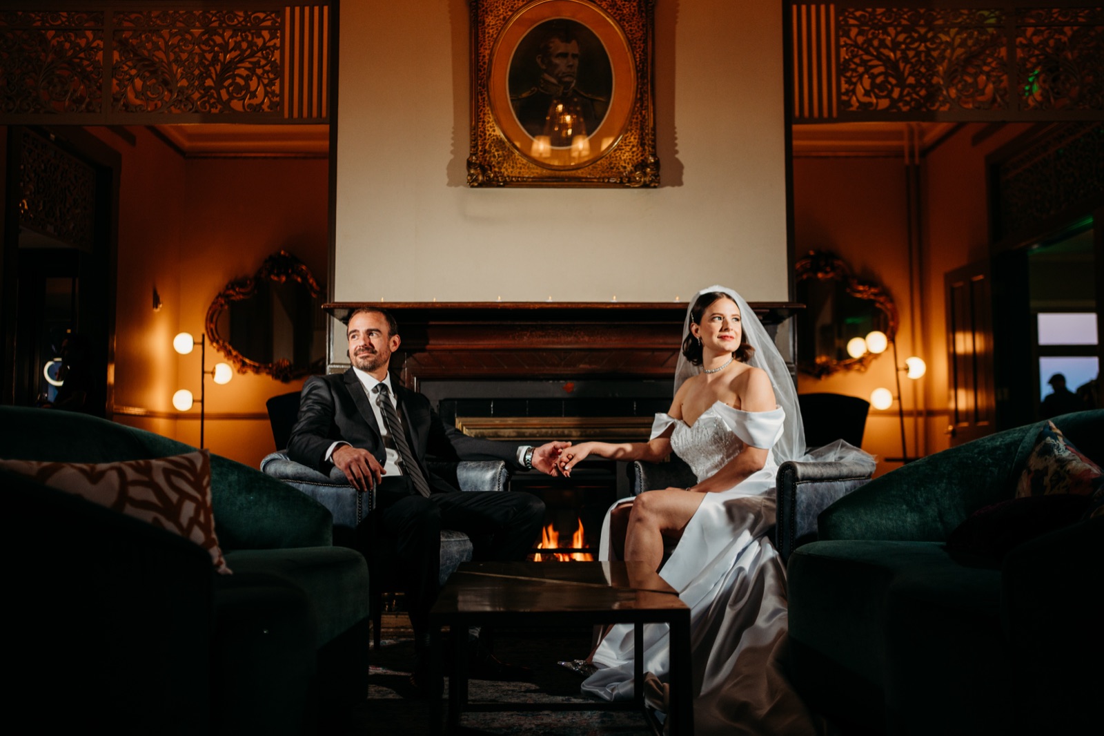 Bride and groom seated in facing chairs beside a fireplace, holding hands, in a candlelit historic lounge